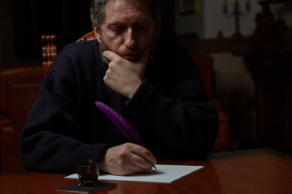 Человек с бородой сидит за большим полированным столом и пишет на белом листе гусиным пером и чернилами