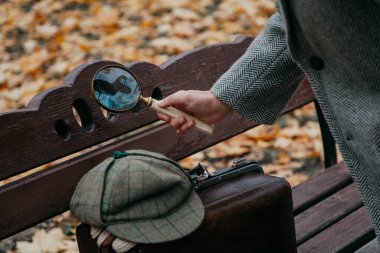 Gri ceketli sağ el, sonbahar parkındaki bir bankın üzerinde büyüteç tutuyor. Yakınlarda deri bir halı çantası var.
