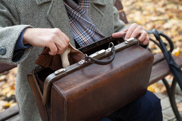 Gri ceketli adam ahşap bir bankta oturuyor. Açık deri bir çantadan bir eldiven çıkardı. Kapat.