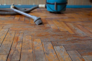 Eski parke zemin kapanıyor. Boya ve vernik ahşap tahtalardan soyuluyor. Arka tarafta mavi bir elektrikli süpürge var.