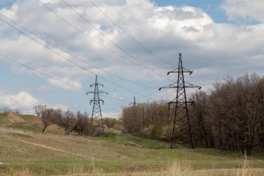 Yeşil çimenli bir tepede duran iki elektrik teli destekçisi. Sağ tarafta büyük ağaçlar var. Gökyüzünde beyaz bulutlar var.