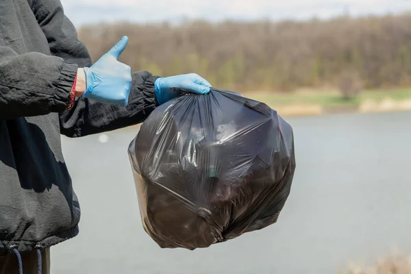 做对的事 生态学家的手戴着蓝色橡胶手套 手里拿着一个装有垃圾的黑色塑料袋在水库岸边 右手有红晕的迹象 — 图库照片