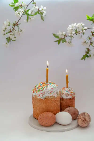 Easter still life. Two Easter cakes and eggs stand on a white background. Burning candles are inserted into the cakes. Top view of a tree branch with white flower