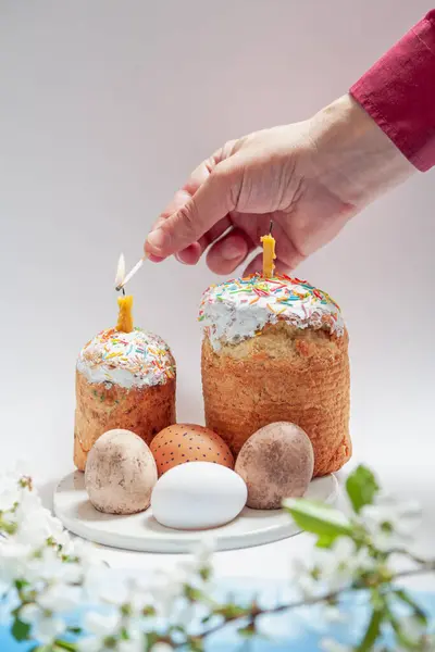 Two Easter cakes and eggs stand on a white background. Candles are inserted into the Easter cakes. A hand lights a candle with a match. Below is a blue cloth and a tree branch with white flower