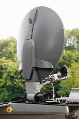 Arka planda ağaçlar olan gri uydu anteni. Canlı üretim aracının üstünde iletişim ekipmanları var. Acil durdurma düğmesi