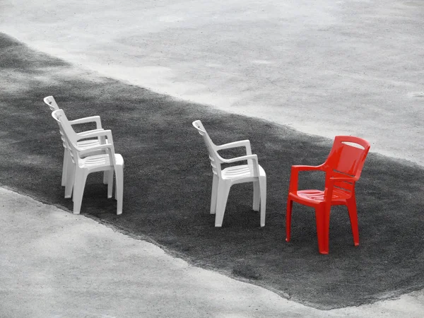 Three white chairs and one distinct red pointing into another direction. Placed on dark asphalt stripe amid lighter one