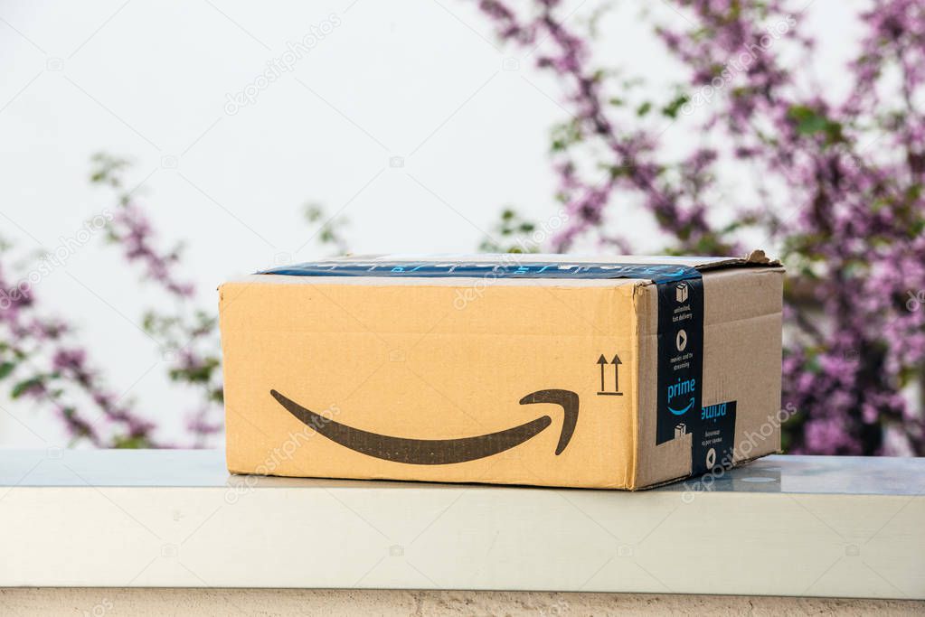 PARIS, FRANCE - APR 19, 2018: Side view of new Amazon Cardboard box left outdoor the house by courier with large Amazon Smile logotype against defocused tree background