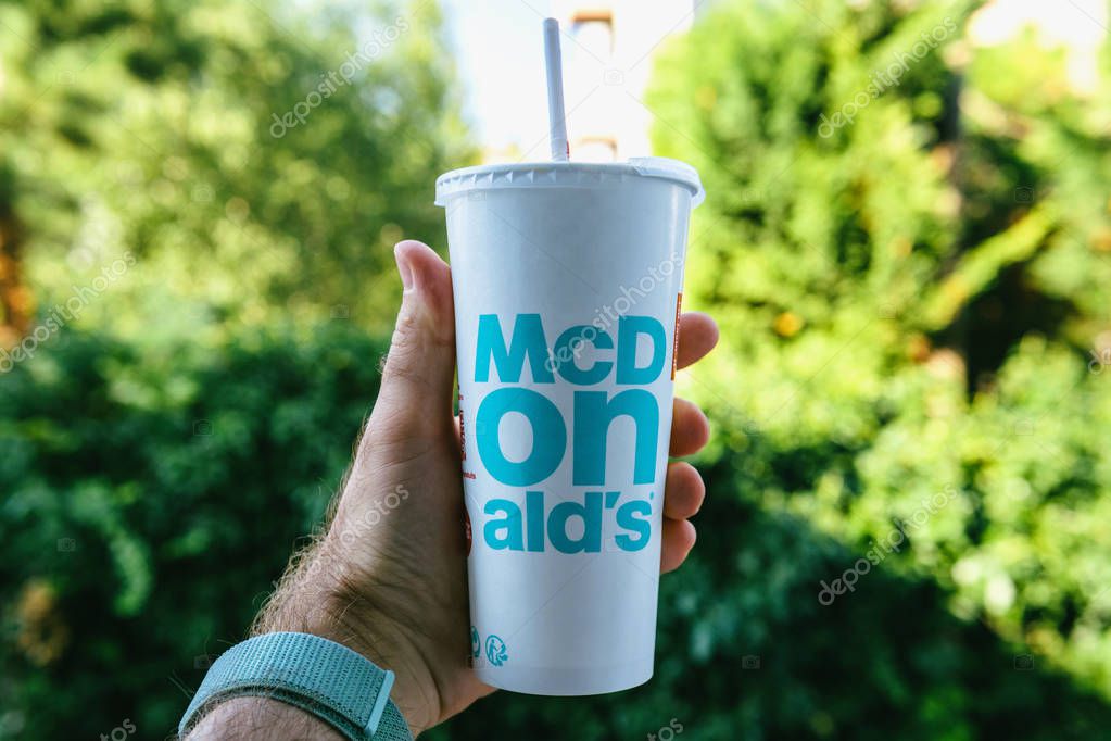 PARIS, FRANCE - JUL 27, 2018: Man holding outdoor against green background a cup of McDonalds Cola with plastic straw with blue text near one of the worlds largest fast food restaurants