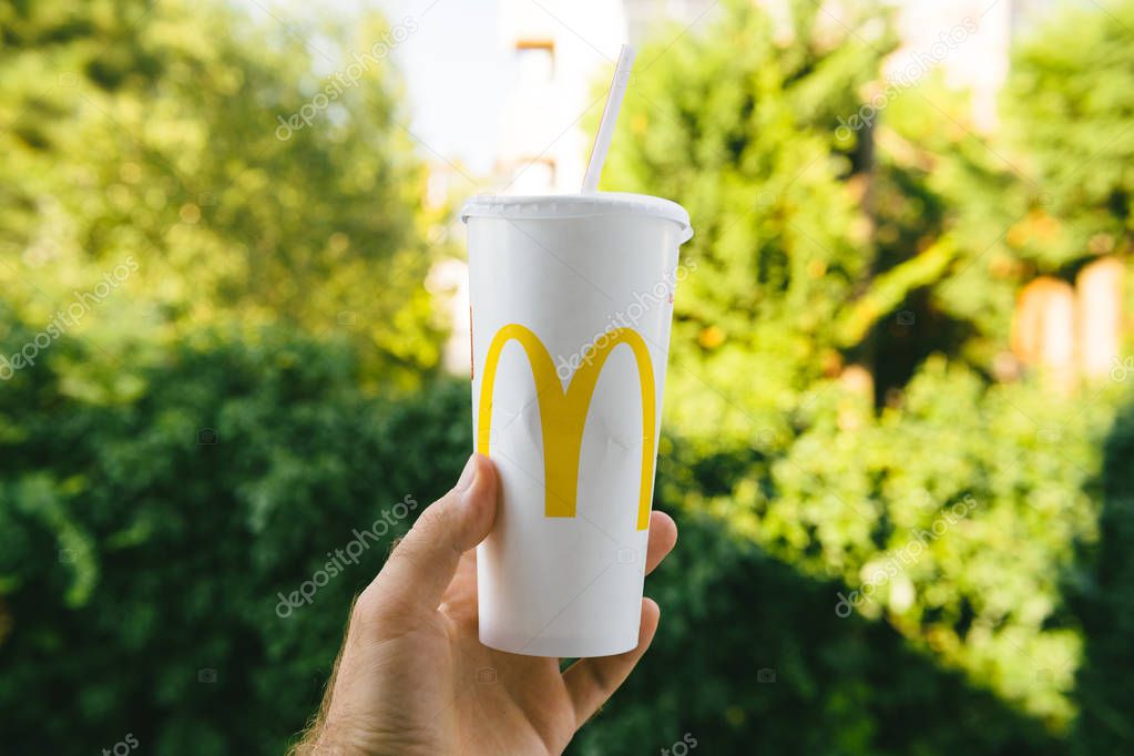 PARIS, FRANCE - JUL 27, 2018: Man holding outdoor against green background a cup of McDonalds Cola with plastic straw with yellow logo near one of the worlds largest fast food restaurants