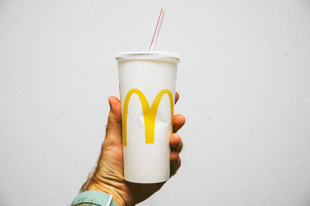 PARIS, FRANCE - JUL 27, 2018: Man holding against white background a cup of McDonalds Cola with plastic straw with yellow logo near one of the worlds largest fast food restaurants