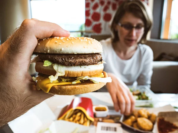 Male hand holding delicious double hamburger woman eating nuggets — Stock Photo, Image