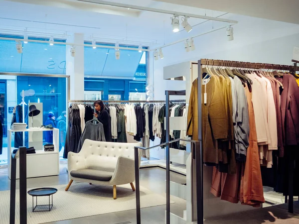 Vista desde la calle de una mujer comprando dentro de una tienda de ropa con múltiples prendas de lujo modernas — Foto de Stock