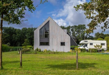 66 numaralı Hollanda konutu ahşap dış görünüşlü ve geniş pencereli, parçalı bulutlu gökyüzünün altında park halindeki bir karavan minibüsünün yanında..