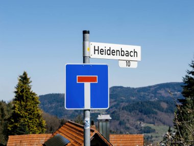 Almanya 'nın Kara Orman bölgesinde açık bir gökyüzü altında çatıların ve ormanlı tepelerin önüne Heidenbach sokak adının yazıldığı mavi bir çıkmaz sokak tabelası..