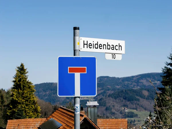 Almanya 'nın Kara Orman bölgesinde açık bir gökyüzü altında çatıların ve ormanlı tepelerin önüne Heidenbach sokak adının yazıldığı mavi bir çıkmaz sokak tabelası..
