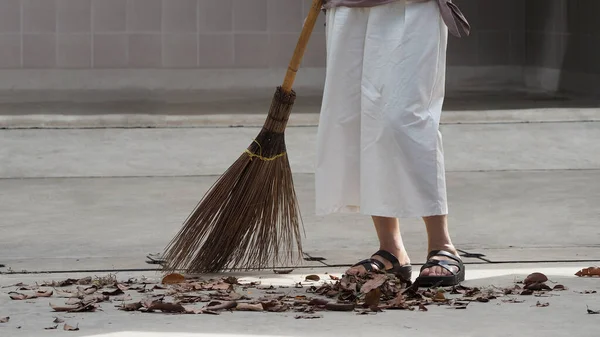 Woman sweeping dry leaves on the cement floor with long wood broom and keeping outdoor clean everyday which images showed middle aged female leg with white pants and black color sandal.