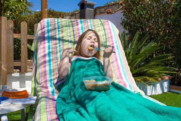 年歳金髪子カラフルな背景の上の緑のタオルでデッキチェアや庭のハンモックに横になっているプラスチック製のトレイからフォークでパスタ マカロニを食べる — ストック写真