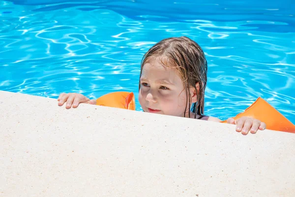 4岁的金发碧眼的孩子 胳膊上有橙色的漂浮物袖子 紧紧抓住或紧紧抓住游泳池的路边 用蓝色透明的水 — 图库照片