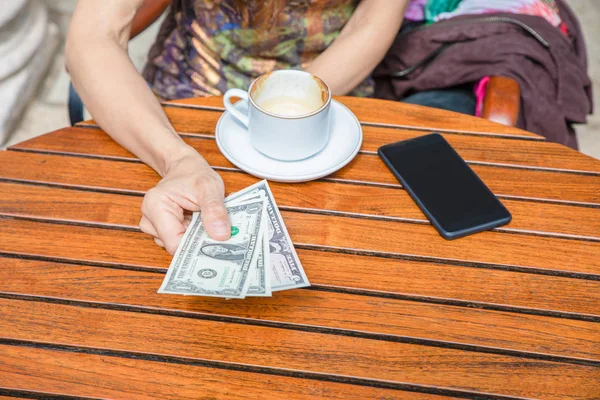 妇女手与美元钞票的细节支付一个卡布奇诺咖啡杯在棕色木桌上 与黑色手机 — 图库照片