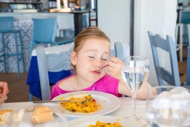 dört yaşında sarışın kız memnuniyeti ifade yüz, restoranda oturan beyaz plaka İspanyol paella pirinç yeme portresi