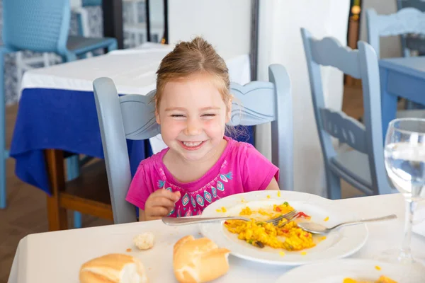 portrait of four years old blonde girl laughing, and eating Spanish paella rice sitting in restaurant