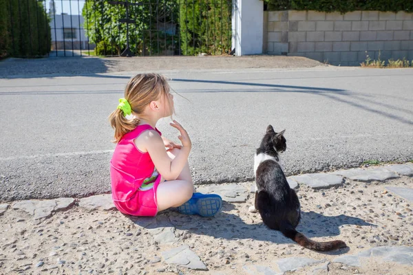funny friends: little girl, five years old child sitting on the ground next to white and black cat, looking at the street together in Summertime