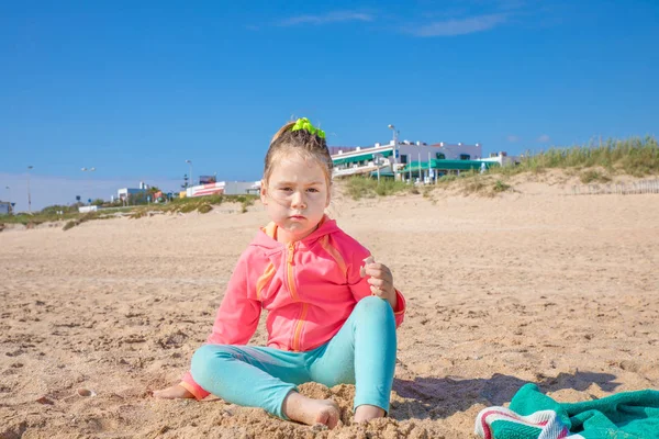 cute four years old blonde girl with pigtail, pink jacket and green leggings, sitting on the sand looking at with challenging expression face, on beach named Palmar, in Vejer, Cadiz, Spain