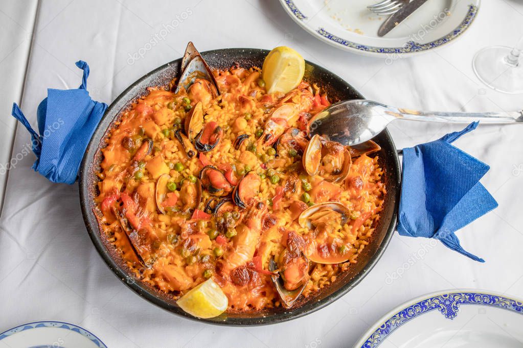 above view of typical Spanish paella with seafood (mussel, prawn, lobster, squid, clam), rice and lemon ready to serve on white tablecloth of restaurant