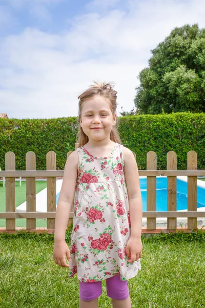 portrait of four years old blonde happy girl with dress, looking at and smiling, standing in a garden next to a swimming pool
