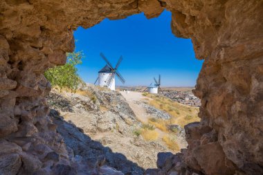 two classic wind mills through a hole in wall in Consuegra town (Toledo, Castilla La Mancha, Spain). Built from the Sixteenth to the Nineteenth century. Typical from Quixote land and his book clipart