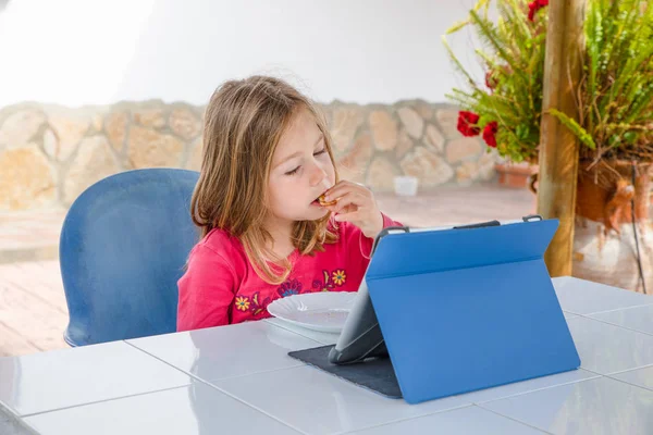 Kleines Mädchen sitzt vor dem digitalen Tablet und isst Kekse — Stockfoto