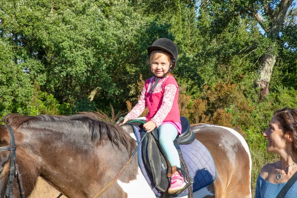 Nettes kleines Mädchen auf einem Pferd, das lächelnd neben seiner Mutter steht Stockbild