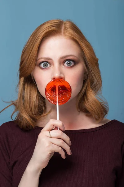 Fashion portrait young woman and lollipop is having fun. Redhead girl closes mouth with a Lollipop, looking in surprise at the camera.