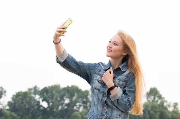 美しい金髪女性長い髪と笑顔でジーンズは自然と空の背景に Selfie を作る — ストック写真