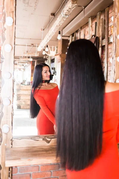 メイクや髪のケア後に鏡で見ている美容サロンで美しいファッショナブルな若い女性 赤いドレスが笑顔で幸せな女の子 — ストック写真
