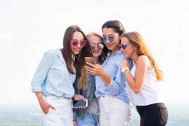 Dört genç kadın güneş gözlüğü smartphone ayakta binanın çatısına kullanın. Dostluk ve modern teknoloji kavramları. Genç öğrenci kız iyi eğlenceler