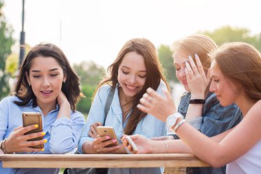 Bir grup genç kız öğrenci Park ve akıllı telefonlar tutan bir şey tartışırken bir gülümseme ile bir masada oturuyor. sohbet, Internet, teknoloji kavramları