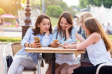 Genç mutlu öğrenci kızlar akıllı telefonlar parkta kullanıyorsunuz. Bir grup bir masada oturan ve konuşmak güzel gülümseyen kadın