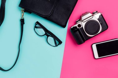 Düz yatıyordu seyahat aksesuarları ile smartphone, Gözlük, siyah çanta ve fotoğraf makinesi renkli mavi ve pembe arka plan üzerinde. Üstten görünüm, kopya alanı