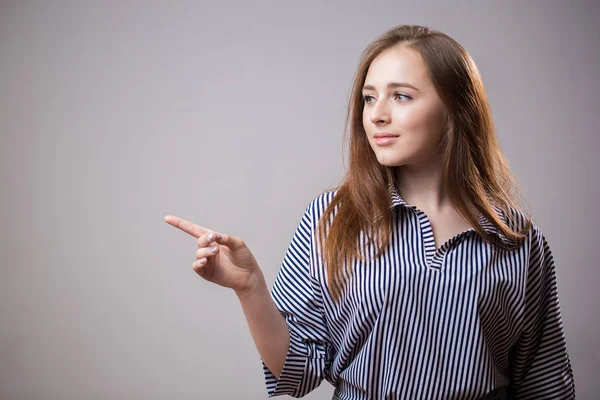 Cute young woman points a finger away on a light gray background with copy space or text, advertising, image. Attractive redhead student girl something advertises, represents a product