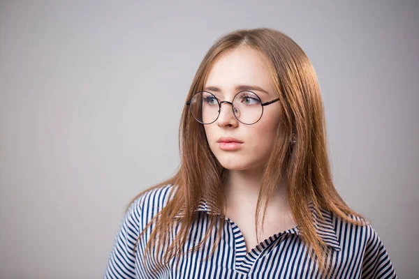 Unhappy, sad young student girl with glasses looking away on grey background with copy space. The woman was offended, upset. Bad mood, emotions, bad day and news, resentment concept