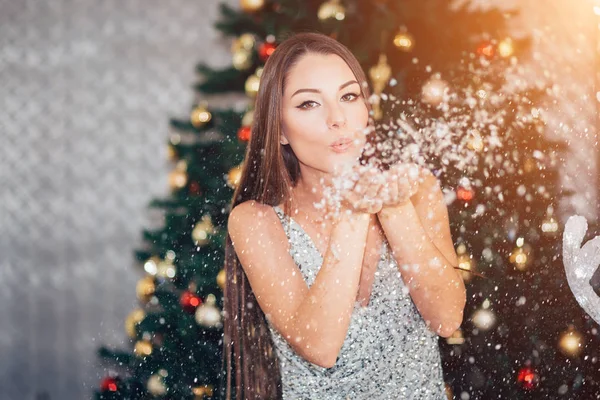 Beautiful brunette woman in new year silver dress blowing snow with hands on Christmas tree background. Holiday, New year, happiness concept