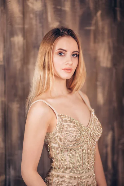 Portrait of elegant beautiful woman in a golden dress on brown wooden background in studio. Blonde girl looking at the camera