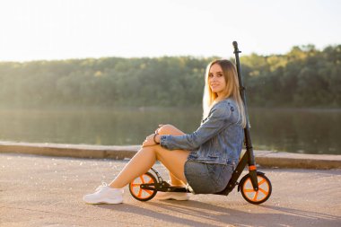 Güzel seksi sarışın kadın denim giysiler içinde onun scooter ve kamera gülümsüyor sokak Rıhtımlar üzerinde oturur. Spor, yürüme, eğlence, eğlence, hobi, yaz konsepti