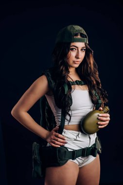 Genç Seksi esmer kız uzun sağlıklı saç askeri elbise ve bir kap ile bir şişe siyah bir arka plan üzerinde tutan ve kameraya benziyor.