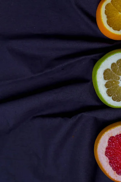 three types of citrus fruits cut pieces of orange grapefruit sweeties half visible on the bottom on a kitchen towel dark grey blue black background with copy space.