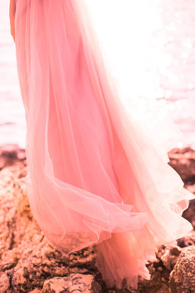 Beautiful girl stands sideways in a pink dress of tulle on the nature of the sea with rocks. The concept of fashion colors 2019, living coral