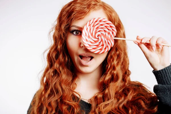 美丽的年轻红发妇女拿着一个红色白色棒棒糖在白色背景 — 图库照片