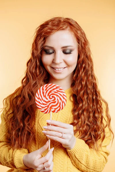 美丽的年轻卷曲红头发的女人拿着一个红色的白色棒棒糖 — 图库照片
