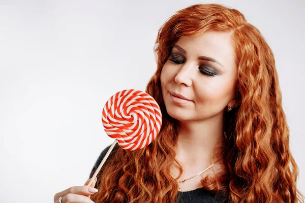 Beautiful curly redhead woman holding a red white lollipop on white background.